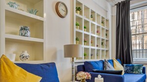Un soggiorno con un divano blu, un tavolino bianco con una lampada e una scaffalatura con oggetti decorativi.