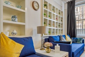 A living room with a blue sofa, a white side table with a lamp, and a shelving unit with decorative items. - 2bed 2bath Apartment Earls Court (London)