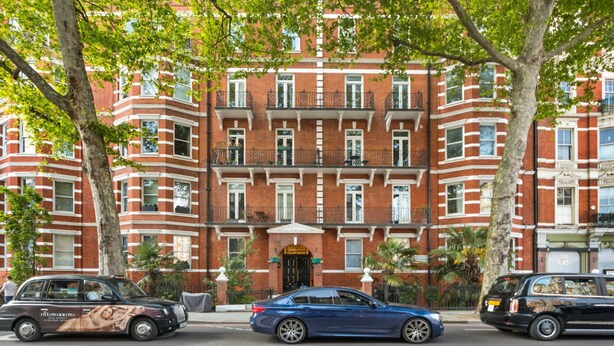 Un edificio in mattoni rossi con numerose finestre e balconi, auto parcheggiate davanti e alberi.