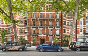 Un edifici de maó vermell amb diverses finestres i balcons, cotxes aparcats a la façana, i arbres.