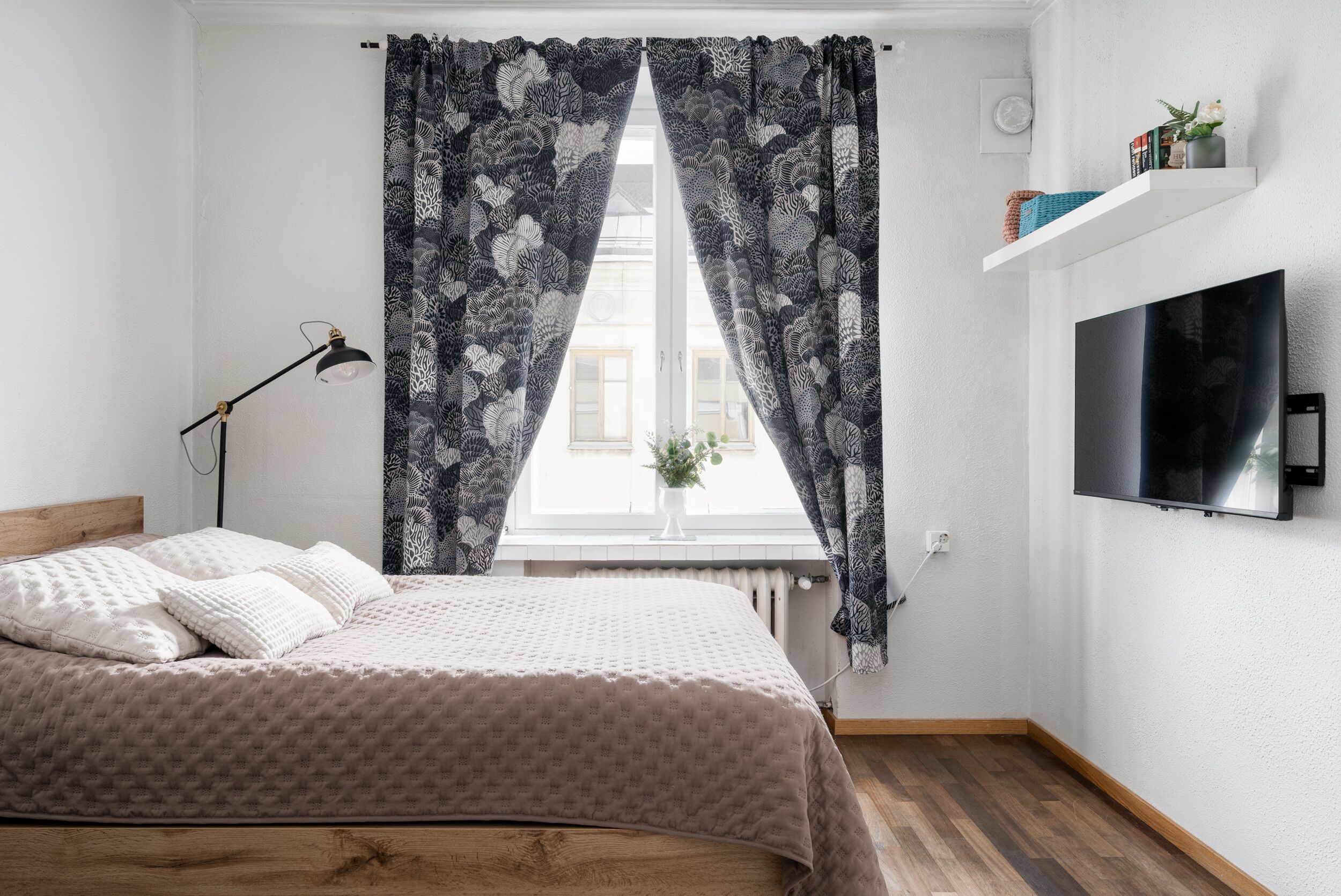 Een slaapkamer met een bed, een raam met behang, een bedlampje, een televisie aan de muur en een plank met spullen.