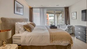A modern bedroom with a large bed, a bedside table, a lamp, a dresser, and a view of the city. - Luxe Creek Riverside Air-conditioned 2br 2bath (London)