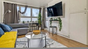 Een moderne woonkamer met een bank, een glazen salontafel, een aan de muur bevestigde televisie en een eethoek met een tafel en stoelen.