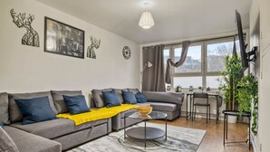 Een moderne woonkamer met een zithoek, een salontafel, een lamp, een plant en uitzicht op de stad door grote ramen.