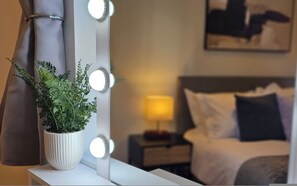 Ein Hotelzimmer mit Schminkspiegel, Beleuchtung, einer Zimmerpflanze, einer Nachttischlampe und einem Bett mit Kissen.