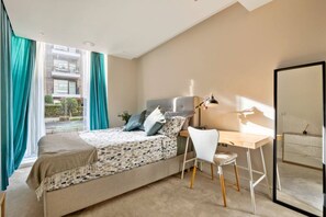 Ein Schlafzimmer mit Bett, Schreibtisch und Stuhl, einem Fenster mit Vorhängen und Blick auf Gebäude.