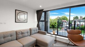 Een moderne woonkamer met een hoekbank, een fauteuil, een klein tafeltje met een plant en een groot raam met uitzicht op een woonstraat.