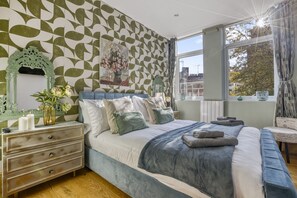 A bedroom with a patterned wall, a large bed with pillows and a blanket, a nightstand with a vase, and a window with curtains. - Elegant 2bed Apt in the Heart of Covent Garden (London)