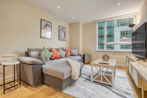 A modern living room with a grey sectional sofa, a small white table, a TV, and a large window with a view of buildings. - 1 Bed 1 Bath Next to Vauxhall Station - Zone 1 (London)