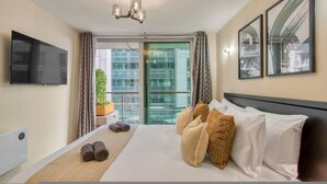 A hotel room with a bed, a flat-screen TV, a balcony with a view of buildings, and a ceiling light fixture. - 1 Bed 1 Bath Next to Vauxhall Station - Zone 1 (London)