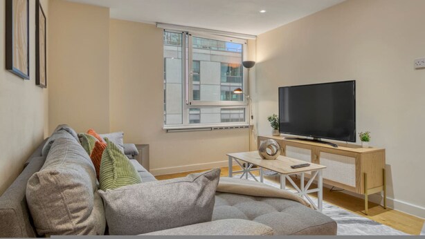 A modern living room with a sofa, a flat-screen TV, a wooden coffee table, and a window with a view of a building. - 1 Bed 1 Bath Next to Vauxhall Station - Zone 1 (London)
