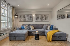 A modern living room with a grey sectional sofa, a round coffee table, and a yellow throw blanket. - Parkside 3 Bed Apartment Hackney (London)