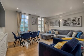A modern living room with a grey sofa, blue chairs, a dining table with chairs, and artwork on the walls. - Parkside 3 Bed Apartment Hackney (London)