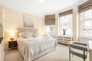 Quarto com cama, abajures, janela com cortinas, uma cadeira e vista para edifícios.