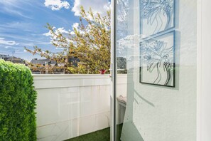 Ein Balkon mit Blick auf Häuser und Bäume, eine weiße Wand mit gerahmten Kunstwerken und ein ordentlich geschnittener Heckenstreifen.