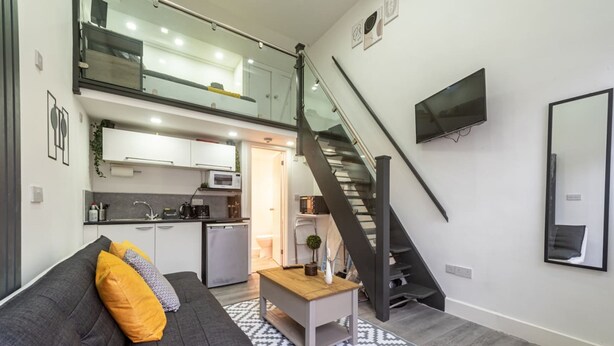 A modern apartment with a loft-style kitchen, a staircase, a flat-screen TV, and a gray sofa with yellow cushions. - Studio in Central Reading- Next to Town Centre (Reading)