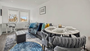 Un salon moderne avec un canapé, une table à manger dressée avec des assiettes et des couverts, un pouf gris et un tapis à motifs.