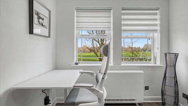 Une pièce avec un bureau blanc, deux fenêtres équipées de stores, un radiateur, une chaise, un tableau encadré et une sculpture en métal noir.