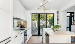 Eine moderne Küche mit weißen Schränken, einer zentralen Kochinsel, einem eingebauten Backofen und einem großen Fenster mit Blick auf einen Garten.
