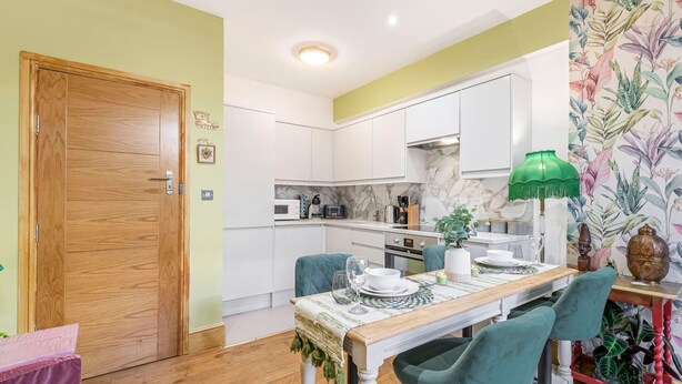 A modern kitchen with white cabinets, a wooden door, and a dining area with a table set for two. - Charming 2bed Garden Apartment in Covent Garden (London)