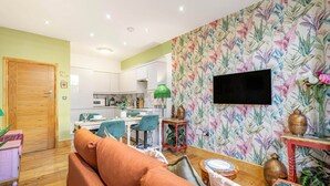 A living room with a sofa, a dining area with chairs, a kitchen in the background, and a TV mounted on the wall. - Charming 2bed Garden Apartment in Covent Garden (London)