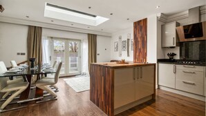 A modern kitchen with a central island, a dining area with a glass table, and a large window with curtains. - Mahogany Heights in Greater London (London)