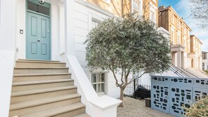 A staircase leading to a white door with a blue frame, a tree in the foreground, and a blue fence with geometric cutouts. - The Grand Camden Garden 5BR Home (London)