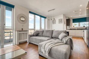 A modern living room with a grey sectional sofa, a white radiator, and a view of the city through large windows. - Modern Charming 2 bed Flat With Amazing Views (London)