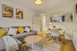 Ein modernes Wohnzimmer mit grauem Sofa, einem runden Couchtisch und einem Essbereich mit kleinem Tisch und Stühlen. An der Wand befinden sich abstrakte Gemälde, ein großer Spiegel und eine Hängeleuchte.