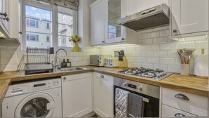 A compact kitchen with a washing machine, a dishwasher, a stove, and a window with a view of buildings. - Comfortable 2-bed in Zone 2 (London)
