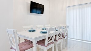 A dining area with a white table and chairs, a flat-screen TV mounted on the wall, and a large window with blinds. - Group-Friendly Home | TV in Every Room, WiFi & Parking (Bon-y-maen)
