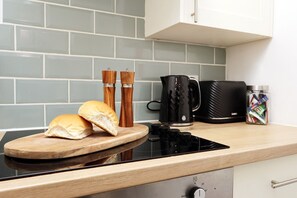A kitchen with a black induction cooktop, a wooden cutting board with bread, two wooden pepper mills, a black coffee maker, and a jar of tea bags. - Studio • Kitchen • Parking • Ideal for Contractors (Gorseinon)