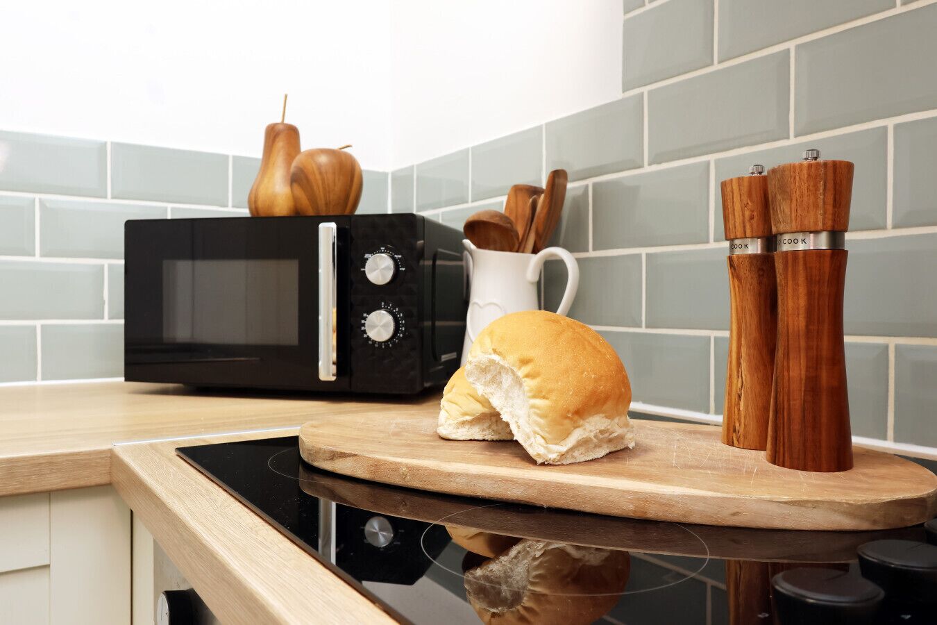 A kitchen with a microwave, wooden pepper shakers, a bread roll, and a white pitcher.