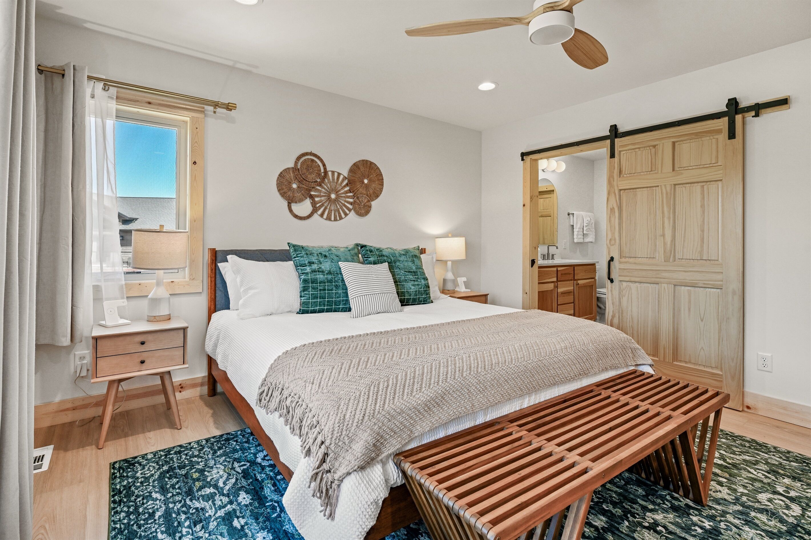 A bedroom with a bed, bedside tables, a bench, a ceiling fan, a window, and a sliding door.