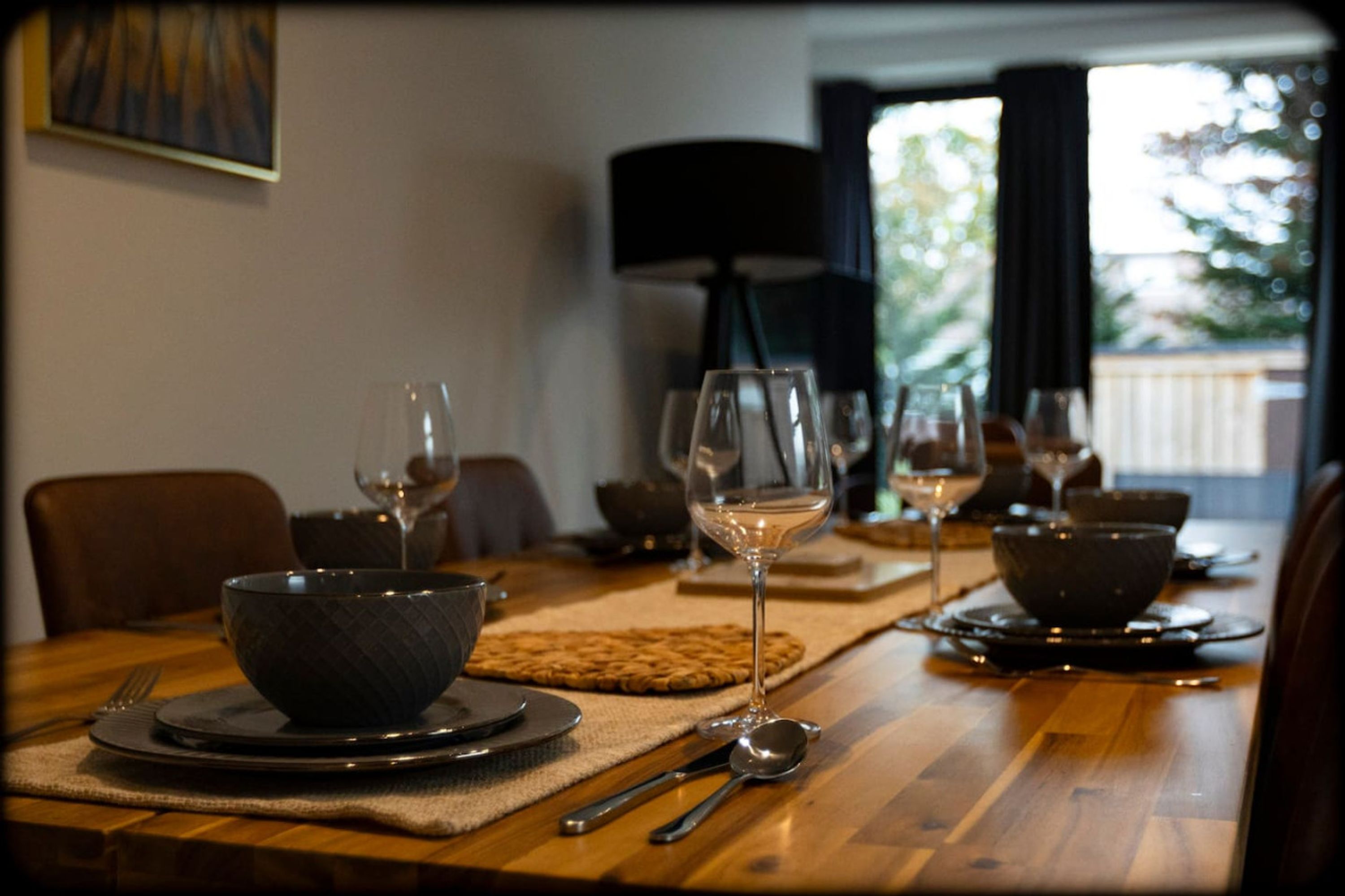 Une table à manger dressée avec des bols, des verres à vin et des couverts, agrémentée d’une lampe et offrant une vue sur l’extérieur.