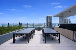 Eine Dachterrasse mit Esstisch und Stühlen, Blick auf das Meer und die Stadtlandschaft, und ein modernes Gebäude im Hintergrund.