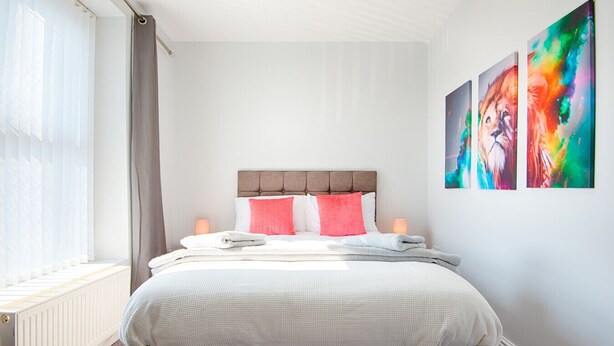 A neatly made bed with a headboard, two bedside lamps, and a window with curtains. - Spacious 3BR Home Sleeps 7 | Parking, WiFi & Near Centre (Plasmarl)