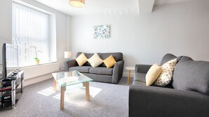 A modern living room with a grey sofa, a glass coffee table, a lamp, and a painting on the wall. - Spacious 3BR Home Sleeps 7 | Parking, WiFi & Near Centre (Plasmarl)