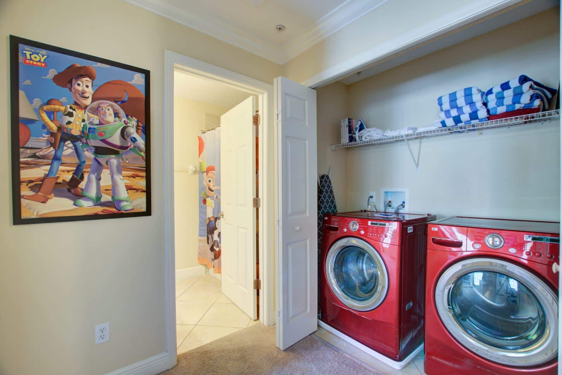 A laundry room with two red washing machines, a shower curtain with a cartoon character, and a shelf with folded towels.