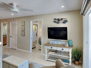 A living room with a flat-screen TV, a ceiling fan, a decorative wall piece, and a sign that reads 'LET'S DO SUNSET'. - Pointe South 504 | Newly Reno, Stunning Gulf Views + Pool (Fort Myers Beach)