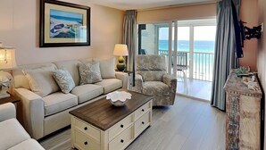 A living room with a sofa, armchair, coffee table, and a painting of a beach scene. - Pointe South 503 | Gulf Views + Heated Pool (Fort Myers Beach)