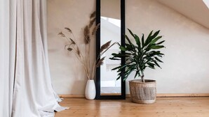 Una habitación con piso de madera, una cortina blanca, un espejo negro alto, un jarrón blanco con plantas secas y una planta en maceta dentro de una cesta tejida.