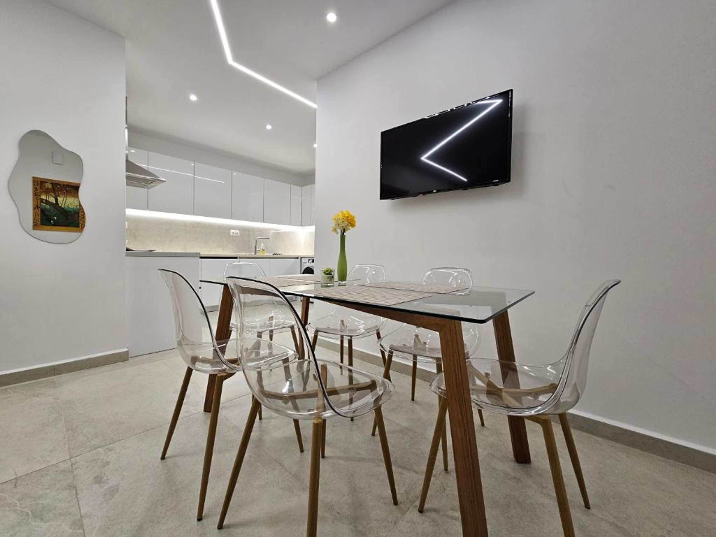 Een moderne eethoek met een glazen tafel en stoelen, een flatscreen televisie aan de muur en een keuken op de achtergrond.