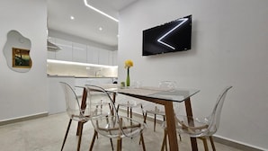 A modern dining area with a glass table and chairs, a flat-screen TV mounted on the wall, and a kitchen in the background. - Sagar Residential Apartment - Athens Town (Nea Ionia)