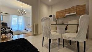 Un salon moderne avec un tapis noir à poils longs, un coin repas avec des chaises blanches, un lustre et une cuisine avec des meubles en bois.