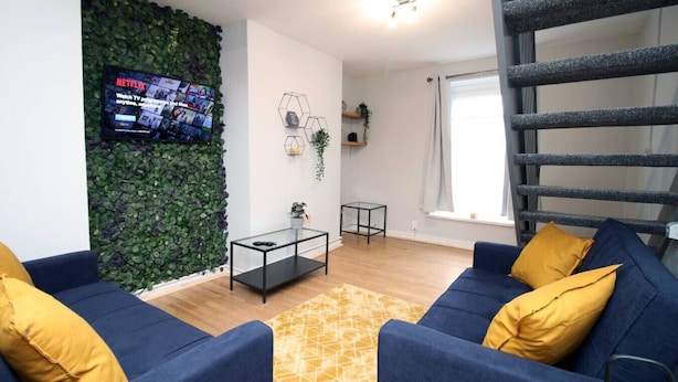 Uma sala de estar com televisão fixada em uma parede verde, uma mesa de centro de vidro, um sofá azul com almofadas amarelas e uma escada.