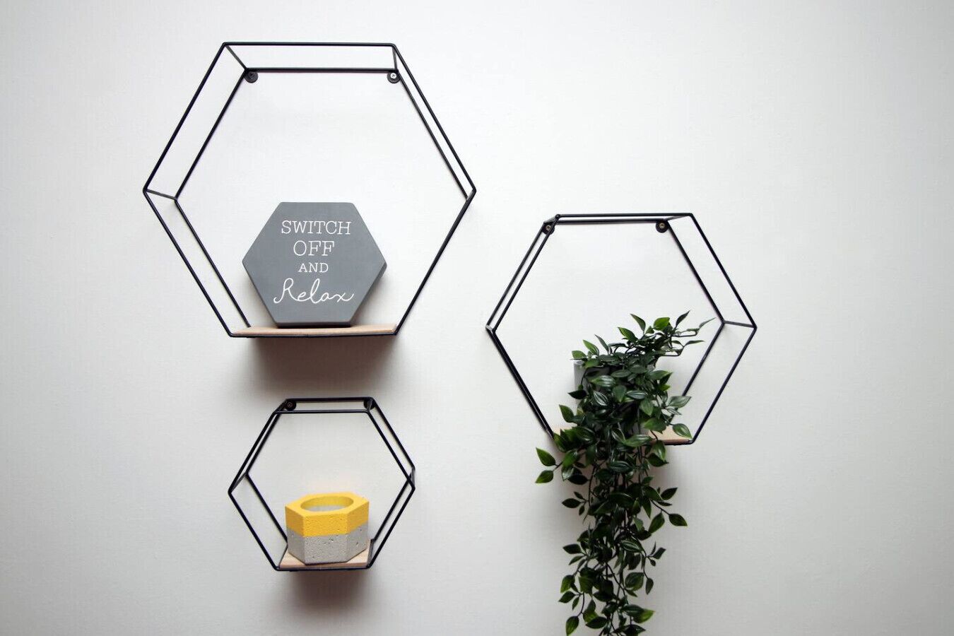 Trois étagères hexagonales fixées au mur, chacune contenant des objets différents : une plaque d’interrupteur, un objet jaune et une plante verte.