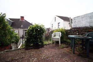 Un jardin à l’arrière d’une maison, avec une terrasse dallée, une chaise en plastique blanche et une table sur laquelle se trouve une assiette.