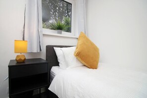 Une chambre à coucher avec un lit, une table de chevet, une lampe, une fenêtre avec une plante et une vue sur les arbres.