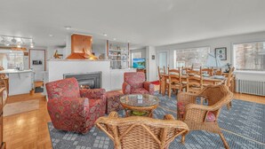 Un salon moderne doté d’une cheminée, de fauteuils à motifs, d’un coin repas avec des meubles en bois et offrant une vue sur la mer grâce à de grandes fenêtres.